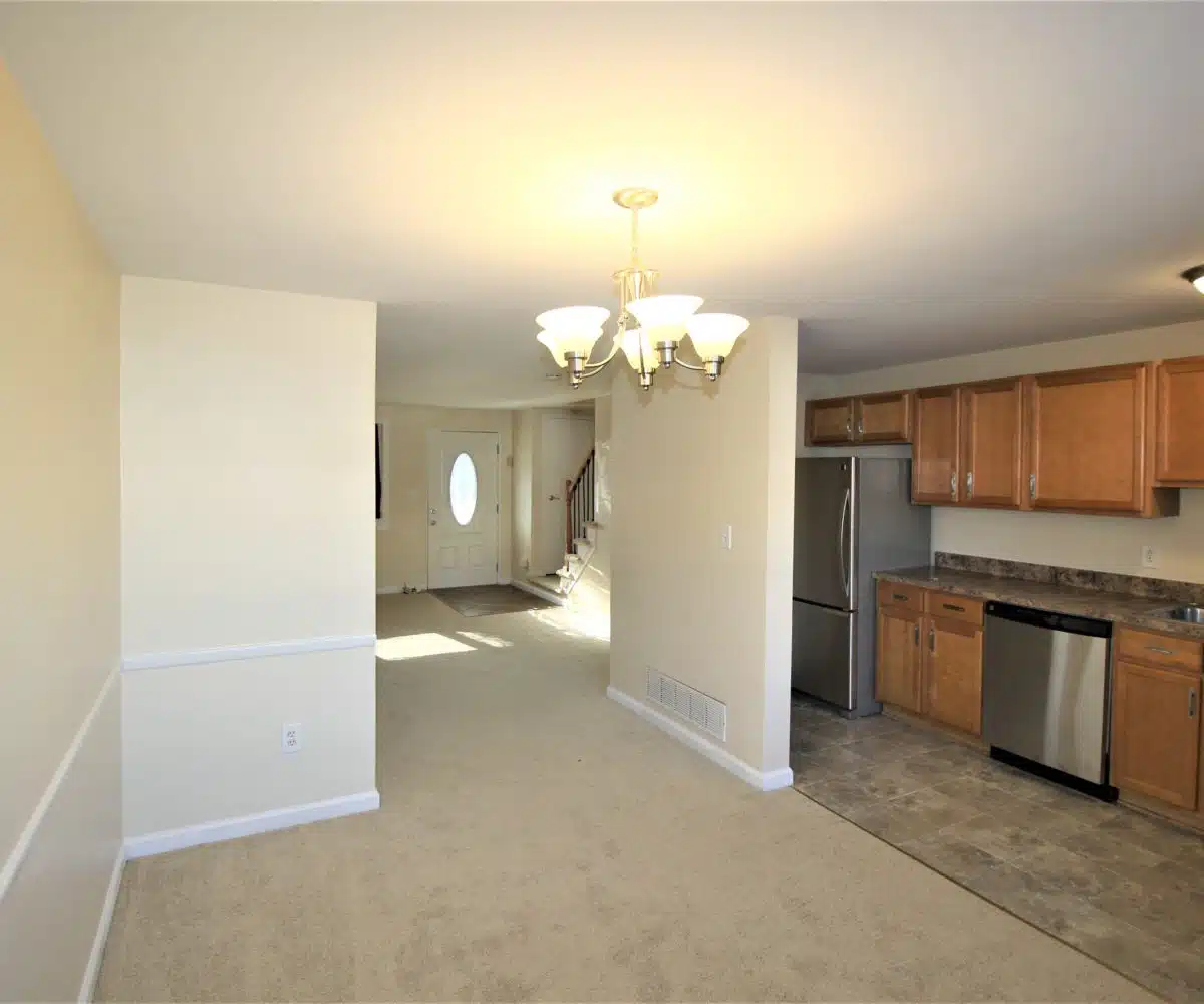 Carpeted living room and dining room with hanging light fixture and kitchen with dishwasher, fridge, wood cabinets and stainless steel sink.