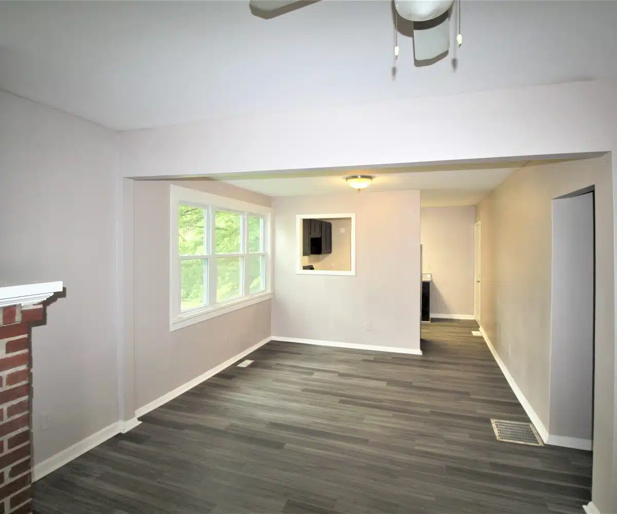 Living room with brick fireplace and dining room with grey flooring, three windows and a ceiling light.