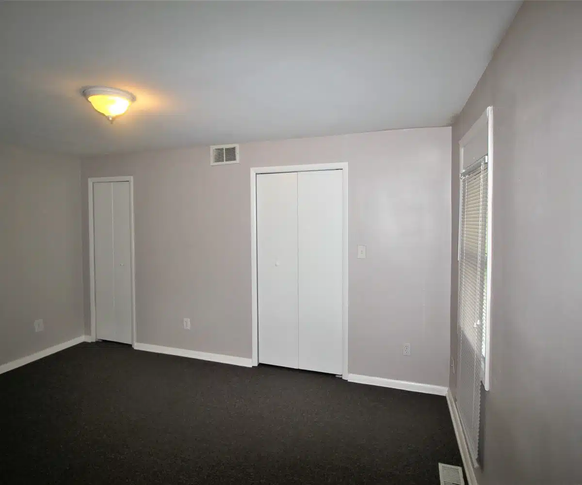 Brown carpeted bedroom with two closets with folding doors a window with a blink and a ceiling light.