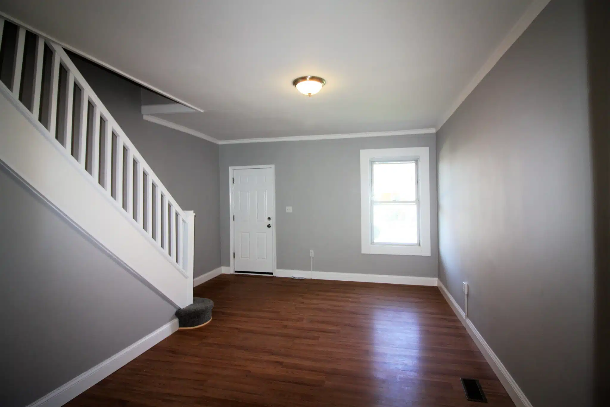 Living room with wood flooring, window, ceiling light and staircase with white hand railing.