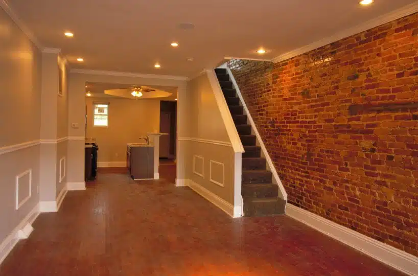 Living room with cherry wood flooring, red brick wall, recessed lighting and brown carpeted stairs.