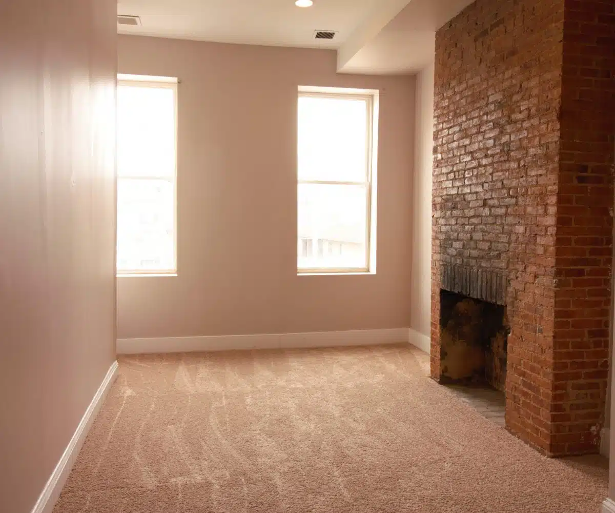 Carpeted living room with two windows and red brick fireplace.