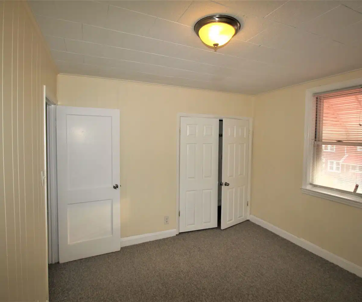 Grey carpeted between with yellow walls, white closet doors and a window with blinds.