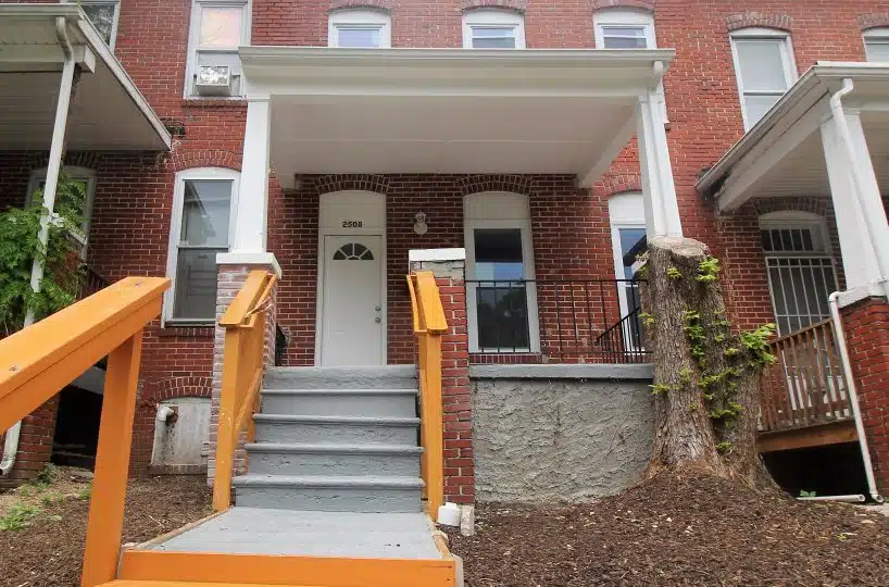 Red brick townhome with orange and grey stairs leading to covered front porch and white front door.