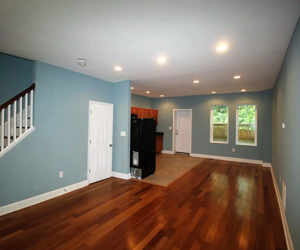 Living room with cherry wood flooring, blue walls, staircase and two windows.