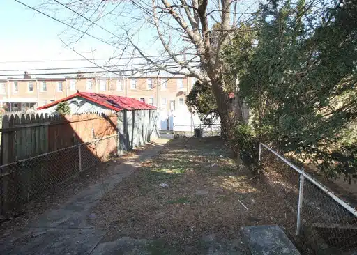Fenced-in backyard with concrete path, leaves, trees and wood fence.