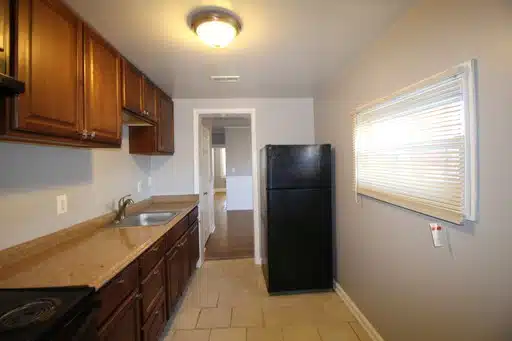 Kitchen with large floor tiles, black fridge, window, granite counters, wood cabinets and a stainless steel sink.