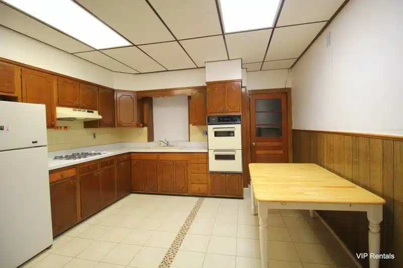 Large kitchen with wood paneling, wood table, built in gas range, double oven and beige floor tiling.