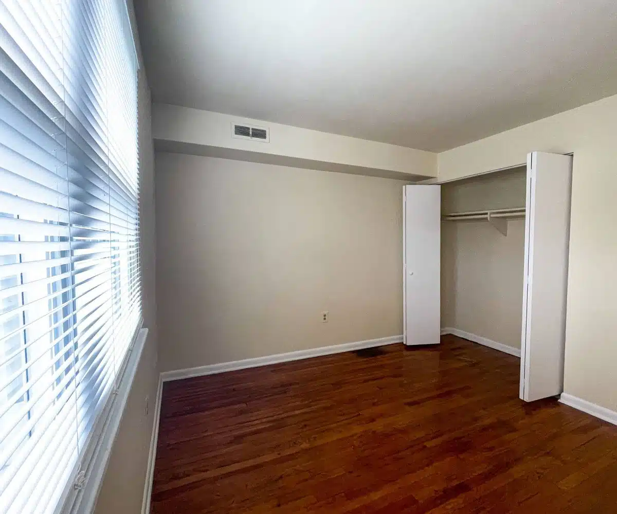An empty bedroom with light colored hardwood floors and a white closet with bifold doors and a window with white blinds.
