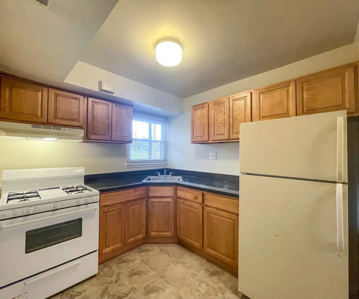 A kitchen with a stove, refrigerator, and sink. There are brown cabinets with black countertops and a window with a blind above the sink.