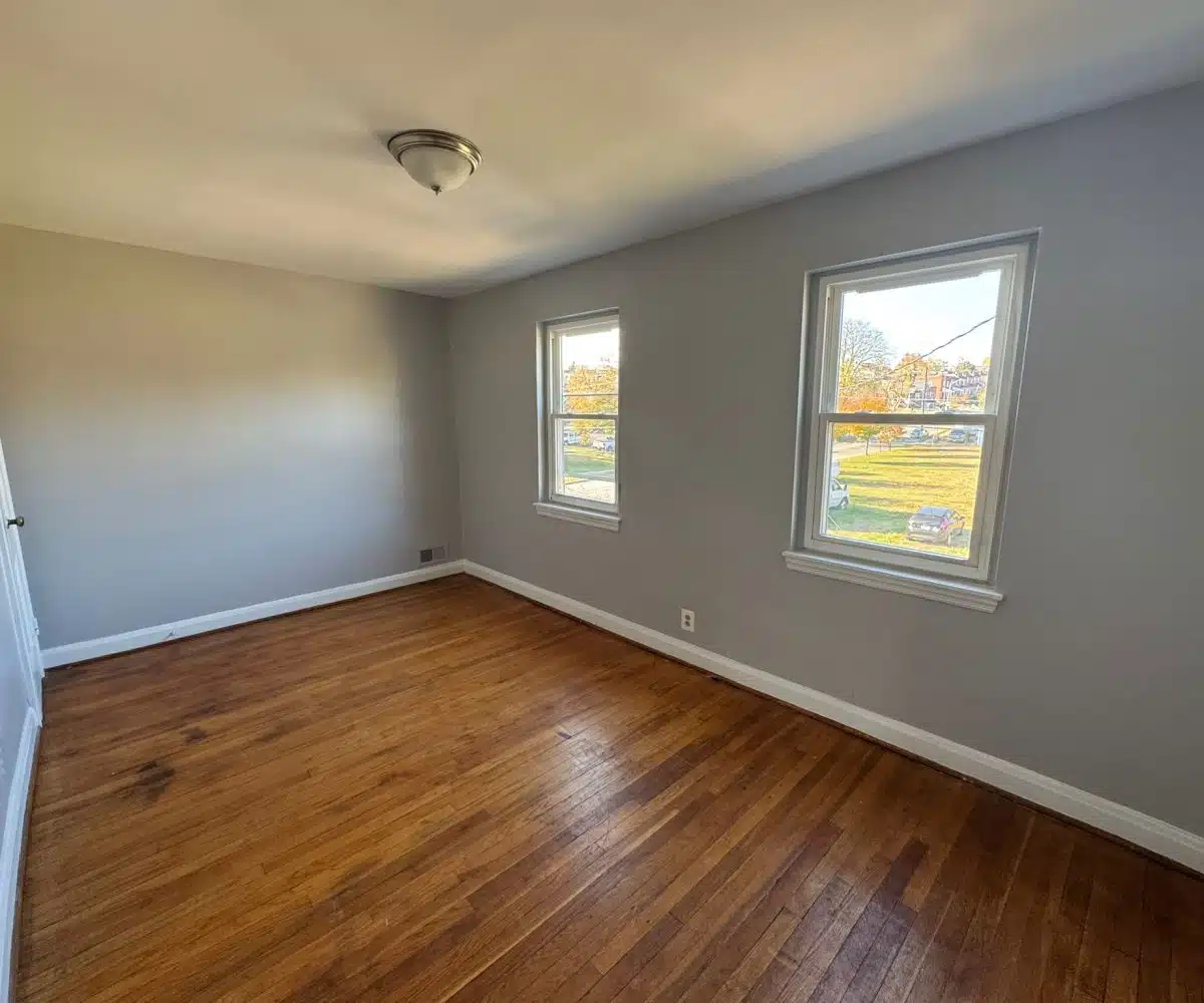 Bedroom with hardwood floors, ceiling light and two windows overlooking grass and trees.