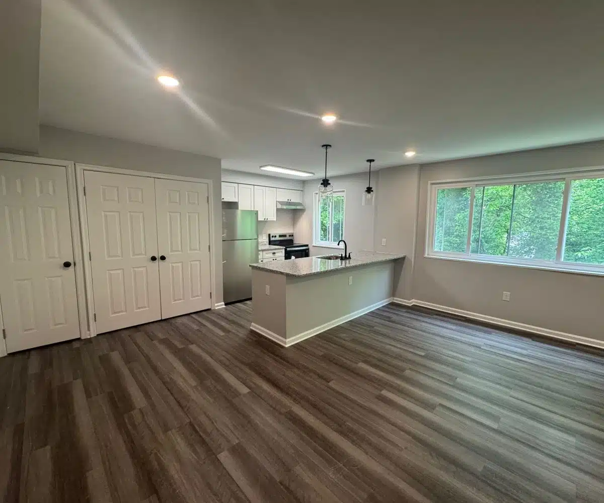 Living room with picture window with view of green trees, ceiling lights and grey flooring.